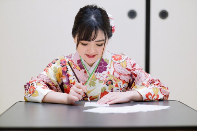 画像：こたつで筆ペンを持ちハガキを書く着物の女性
