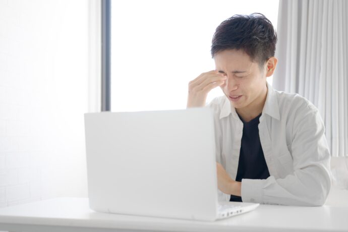 画像：パソコンの前で両目を固く閉じ額に手をやる男性