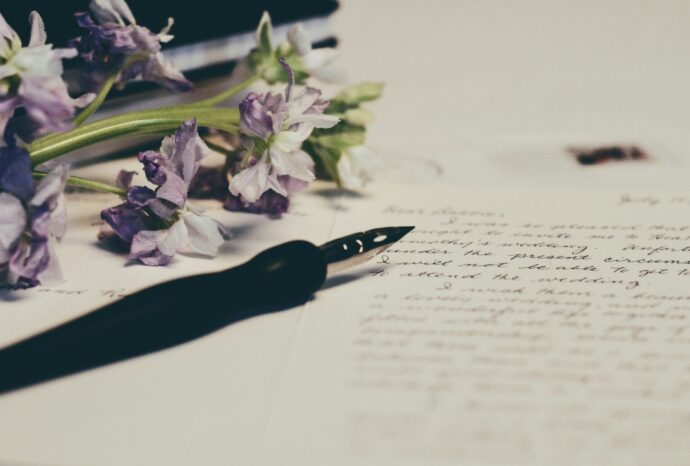 画像：英文が書かれたノートと万年筆と紫色の花