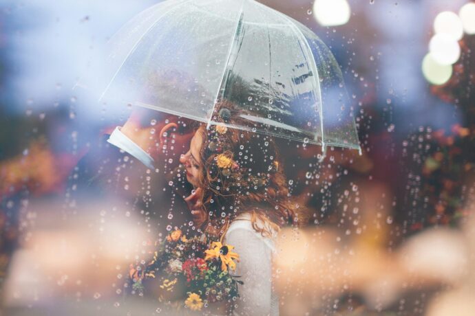 画像：1つの傘を差し笑いあうカップルに雨が降り注いでいるウエディングフォト