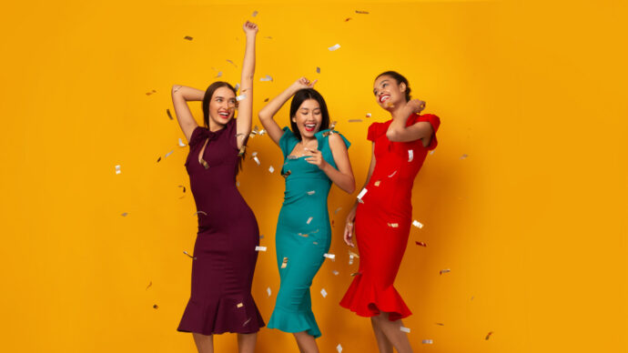 画像：黄色の壁を背景にはしゃいだポーズを取る派手な色合いのドレスを着た女性3人