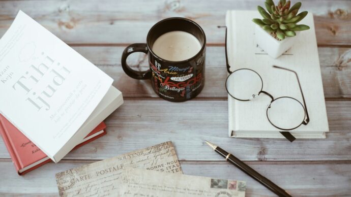 画像：白いウッド調の机の上に並べられたマグカップ、冊子、ペン、眼鏡、観葉植物