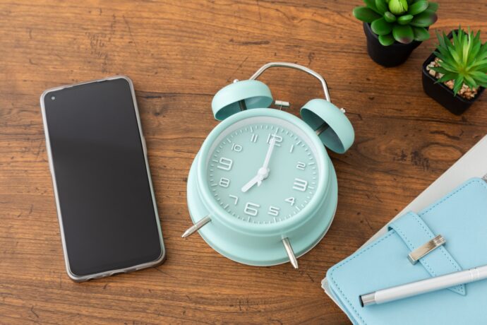 画像：机の上に置かれた水色の目覚まし時計とスマートフォン、手帳と観葉植物