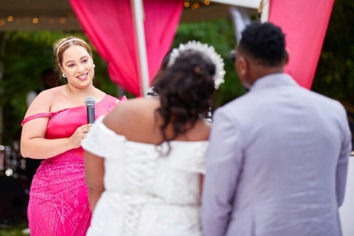 画像：マイクを持つピンクのドレスの女性と肩を寄せ合う新郎新婦